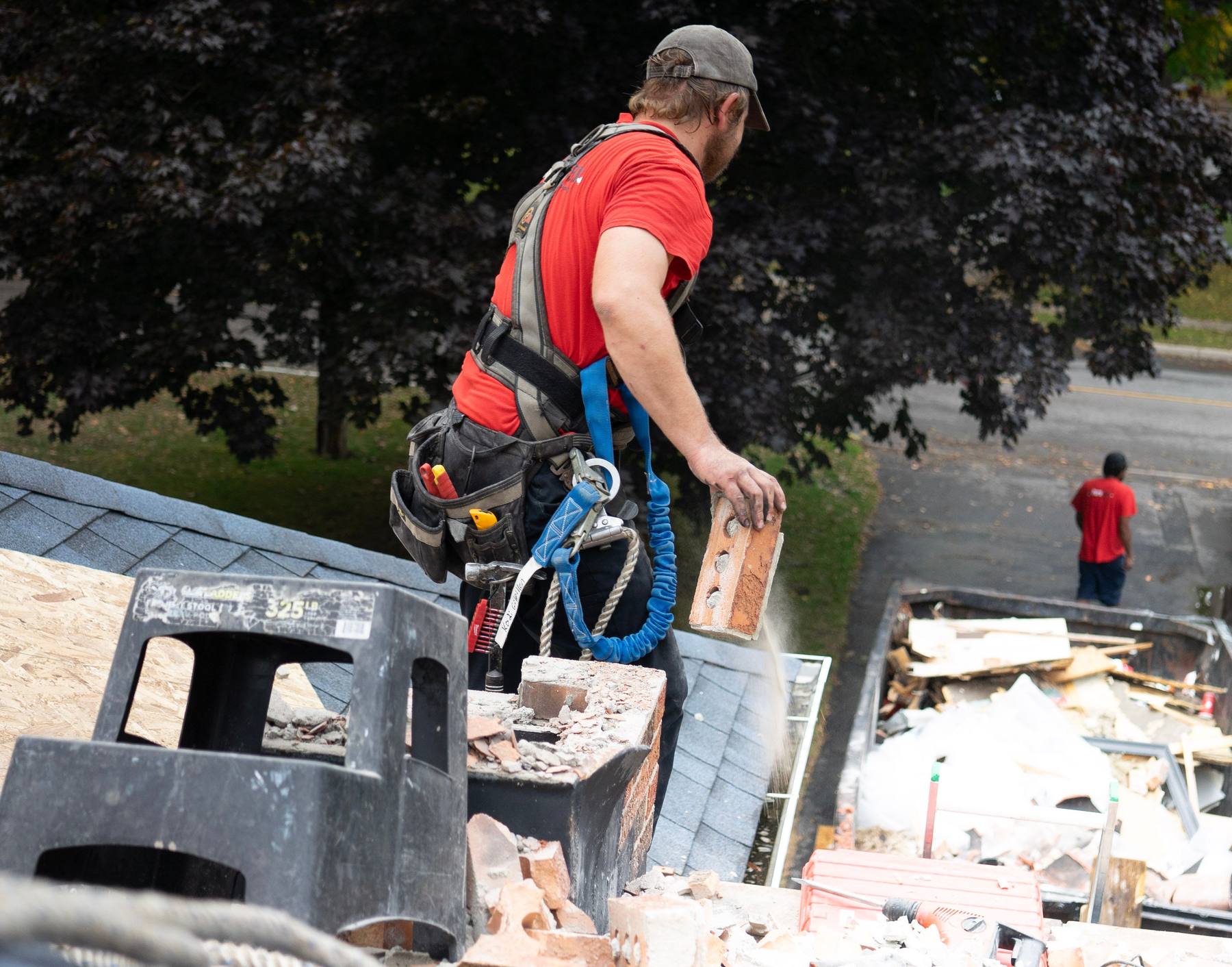 A worker in a safety harness on a residential roof, lifting brick clear of a chimney tear-out