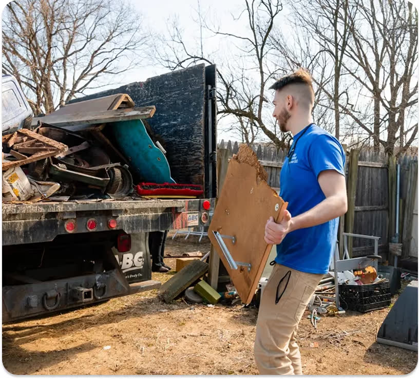 A man in a blue company T-shirt carrying a large wood panel past a dump truck already loaded with debris