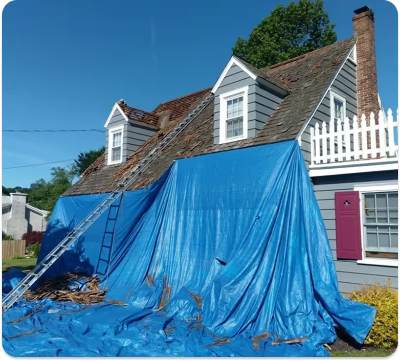 A grey cape-style house mid roof tear-off with blue tarps down one side and a ladder against the eaves